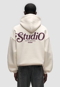 Felpa crema con un grande logo viola "Studio" sulla schiena, caratterizzata da una vestibilità rilassata, cappuccio con cordino e materiale morbido.