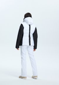 Pufferjack met een zwart-wit kleurblokdesign, voorzien van een capuchon, gewatteerde textuur en gedragen met witte broek en beige laarzen.