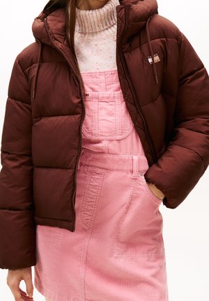 Person iført en mørkebrun dunjakke over en cremefarvet, plettet rullekravetrøje og en lyserød fløjlsnederdel med seler, med hånden i lommen.