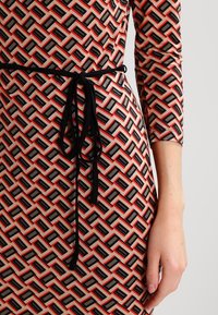 Sukienka w wzór geometryczny w kolorach koralowym, czarnym i beżowym. Posiada czarny pasek wiązany w talii oraz rękawy o długości do połowy ramienia.