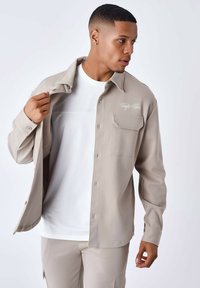 Lys beige skjorte med knapper og en tekstureret stof, med en brystlomme og broderet logo. Båret over en hvid t-shirt med rund hals.