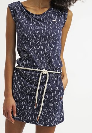 Kobieta ubrana w granatową sukienkę bez rękawów z białymi wzorami morskich koników i białym paskiem ze sznura związanym w pasie, z jedną ręką w kieszeni.