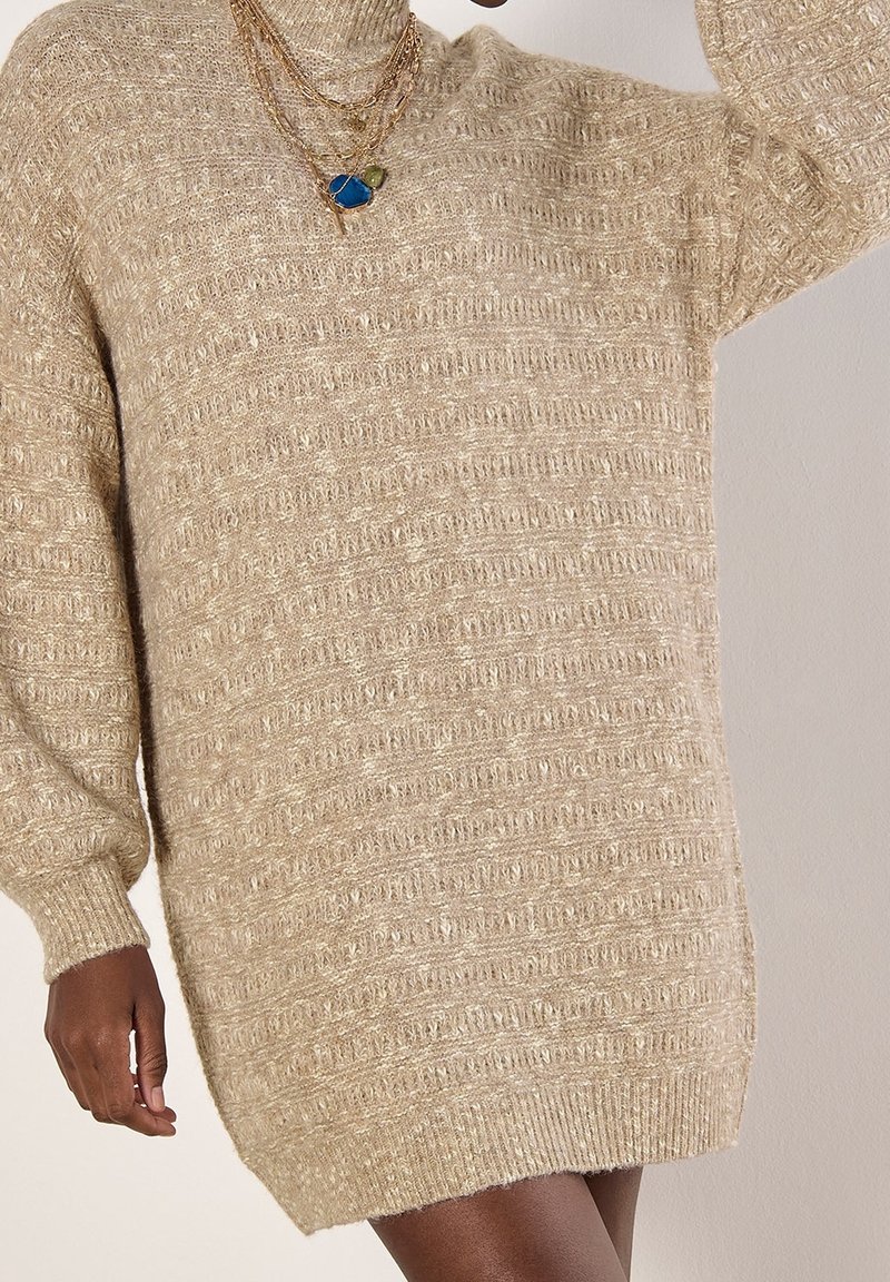 Robe pull en maille texturée beige à manches longues, portée par une personne avec des colliers en or superposés avec des pendentifs bleus et verts.