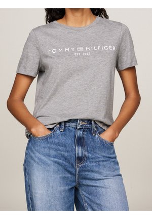 Kobieta w szarej koszulce Tommy Hilfiger z logo i niebieskich dżinsach, ręce w kieszeniach, na jednolitym jasnym tle.