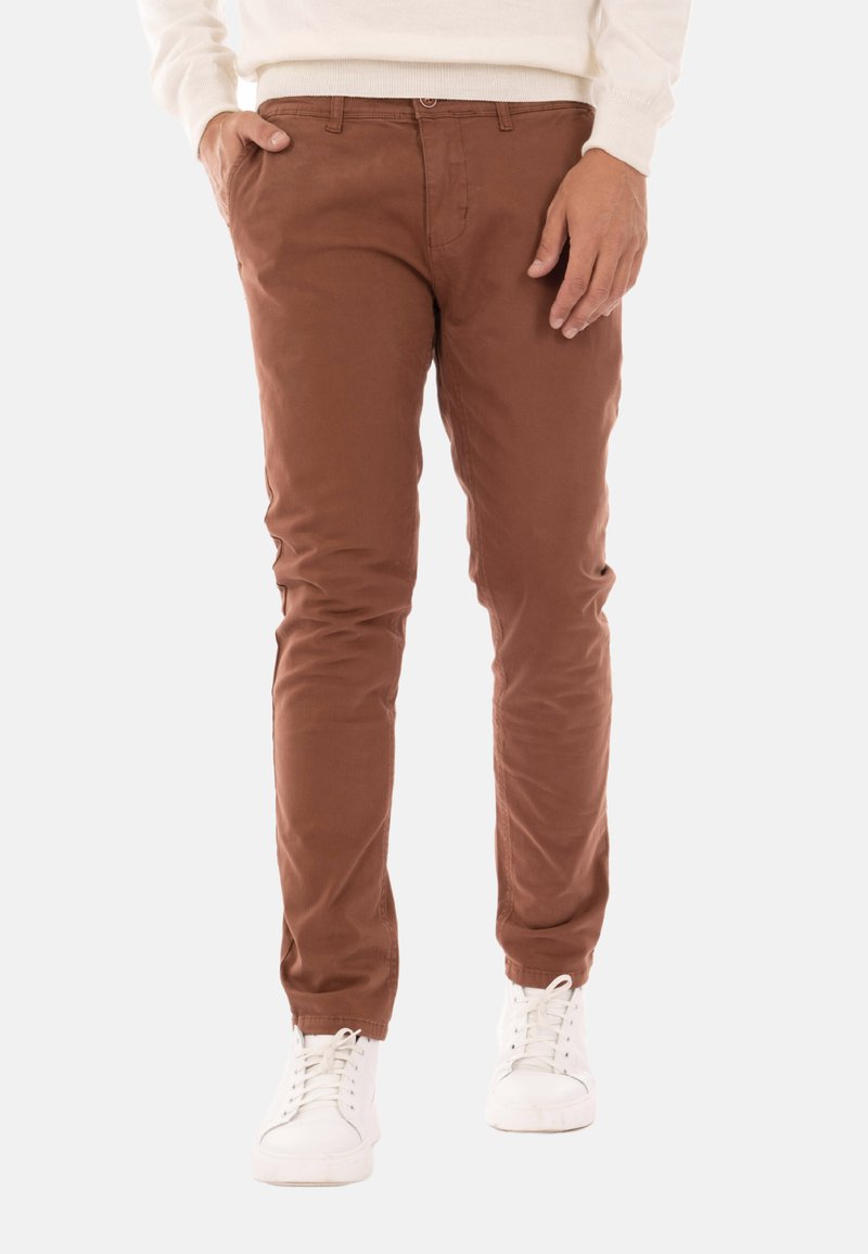 Chinos in cotone marrone con vestibilità slim, tasche frontali e chiusura con zip. Indossati con sneakers bianche a bassa cima. Texture liscia.