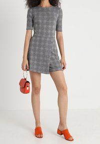 Mono de cuadro gris con mangas cortas y diseño envolvente. Combinado con sandalias de slide naranjas y un pequeño bolso rojo. Textura de tela suave.