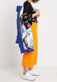 Abrigo acolchado azul con un forro impreso colorido, llevado sobre una blusa negra con flores y pantalones cortos naranjas. Zapatillas blancas completan el conjunto.