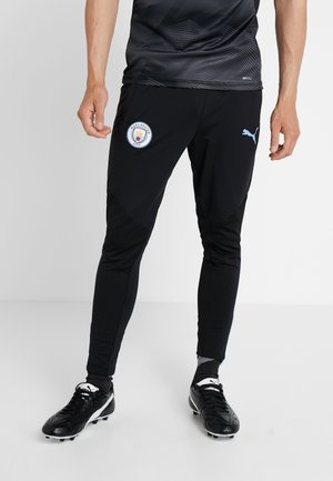 Person, der schwarze Manchester City Jogginghosen mit Puma-Logo und schwarze Fußballschuhe trägt, steht auf einem weißen Boden vor einer schlichten weißen Wand.