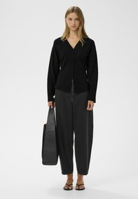 Melnā kardigāna ar V formas izgriezumu, garām piedurknēm un piecām pogām. Kombinēts ar brīviem melniem džinsiem. Modelis tur lielu melnu somu.