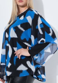Poncho top nero e blu con maniche lunghe, caratterizzato da forme astratte e una texture di tessuto leggero e trasparente.