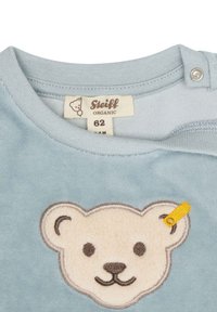 Lichtblauw bovenstuk van biologisch katoen met borduursel van een teddybeer gezicht, zachte textuur, met een klein geel labelaccent nabij het oor. Maat 62.
