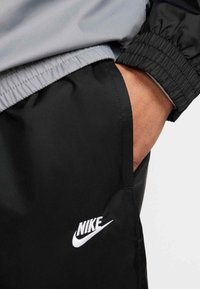 Αθλητικό παντελόνι Nike από μαύρο, λεία ύφασμα, με ελαστική μέση και λευκό λογότυπο στο αριστερό μηρό. Ορατή γκρι επάνω άκρη.