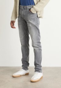 Jeans de denim gris con un corte ajustado, ligeramente deslavados y un diseño clásico de cinco bolsillos. Llevados con zapatillas blancas y una camisa azul debajo de un cárdigan de punto.