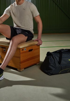 Muž v béžovém tričku a černých šortkách sedí na dřevěném gymnastickém bedeně vedle černé sportovní tašky na podlaze tělocvičny.