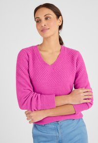 Maglioncino rosa a maglia con scollo a V, motivo a diamante testurizzato sul petto e polsini a coste. Indossato con jeans in denim azzurri chiari.