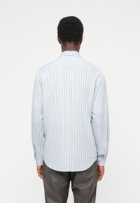 Άνδρας γυρισμένος με την πλάτη, φοράει μακρυμάνικο λευκό πουκάμισο με λεπτές κάθετες ρίγες και σκούρο παντελόνι, πάνω σε απλό λευκό φόντο.