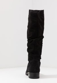 Wysoki czarny zamszowy but z niskim, podpartym obcasem, lekko opadającą cholewą, prezentowany z tyłu na białej powierzchni i tle.