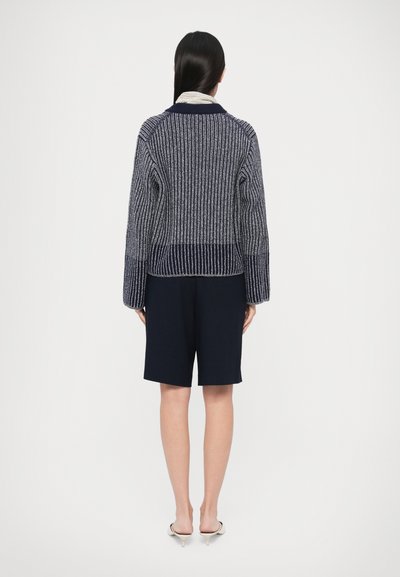 Tmavomodrý rebrovaný pletený sveter s vysokým golierom, detailom na zadnej časti a širokými rukávmi, skombinovaný s tmavými šortkami a nazúvacími topánkami.