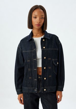 Chaqueta de mezclilla oscura con un corte relajado, gran bolsillo en el pecho, herrajes dorados y detalles de costura en contraste. Presenta un cuello con solapas y cierre de botón.