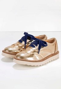 Oxford in oro metallico con lacci blu navy, caratterizzati da un design a punte alate testurizzato, suola platform bianca a contrasto e cuciture decorative.