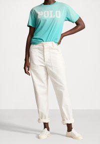 Tyrkysové tričko s krátkým rukávem s potiskem "POLO", kombinované s smetanovými kalhotami s vysokým pasem a bílými teniskami. Neformální design outfitu.