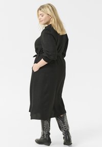 Black, long-sleeve dress with a textured fabric, waist tie, and side pockets. Paired with black cowboy boots featuring silver detailing.