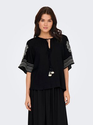 Woman wearing a black blouse with white embroidered patterns on sleeves and tassel ties at neckline, paired with a black skirt.