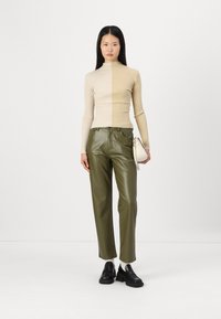 Pull en maille côtelée beige, pantalon en cuir vert olive et chaussures à plateforme noires. Le mannequin tient un petit sac à main blanc. Fond simple.
