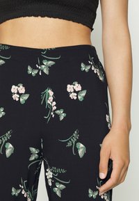 Žena v černých kalhotách se zeleným motýlkem a bílými květinovými vzory, doplněná černým strukturovaným krátkým topem, pravá ruka uvolněně po straně.