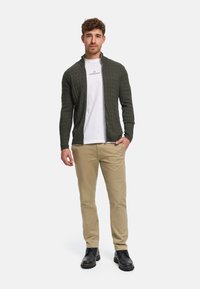 Grön kabelstickad zip-up tröja över en vit skjorta, kombinerad med beige byxor och svarta stövlar. Fokus på textur och kontrasterande färger.