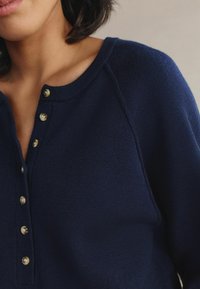 Navy blue knitted pullover with a round neckline and five button placket. Features raglan sleeves and a soft, textured fabric.