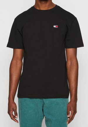 Mężczyzna w czarnej koszulce z krótkim rękawem z małym logo Tommy Jeans na piersi oraz turkusowych sztruksowych spodniach.