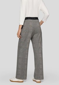 Person trägt graue, karierte Hosen mit weitem Bein, einem schwarzen elastischen Bund und weißen Turnschuhen, steht mit einer Hand in der Hüfte.