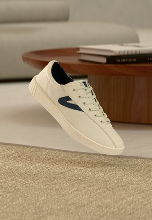 Baskets blanches avec des détails bleu marine flottant au-dessus d'un tapis beige texturé, avec une table basse en bois et des livres en arrière-plan.