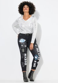 Top plateado de manga larga con escote en V. Jeans negros con parches y estampados gráficos, incluyendo "QUEEN" y "ALL EYES ON ME", con estrellas y diseños de ojos.
