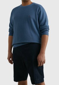 Blauer strukturierter Pullover mit Rundhalsausschnitt, versehen mit einem kleinen Logo auf der Brust, kombiniert mit dunklen Shorts. Der Stoff hat ein geripptes Muster.