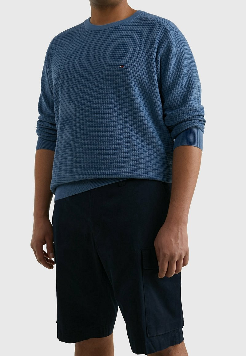 Blauwe gebreide trui met een ronde hals, voorzien van een klein logo op de borst, gecombineerd met donkere shorts. De stof heeft een geribbelpatroon.
