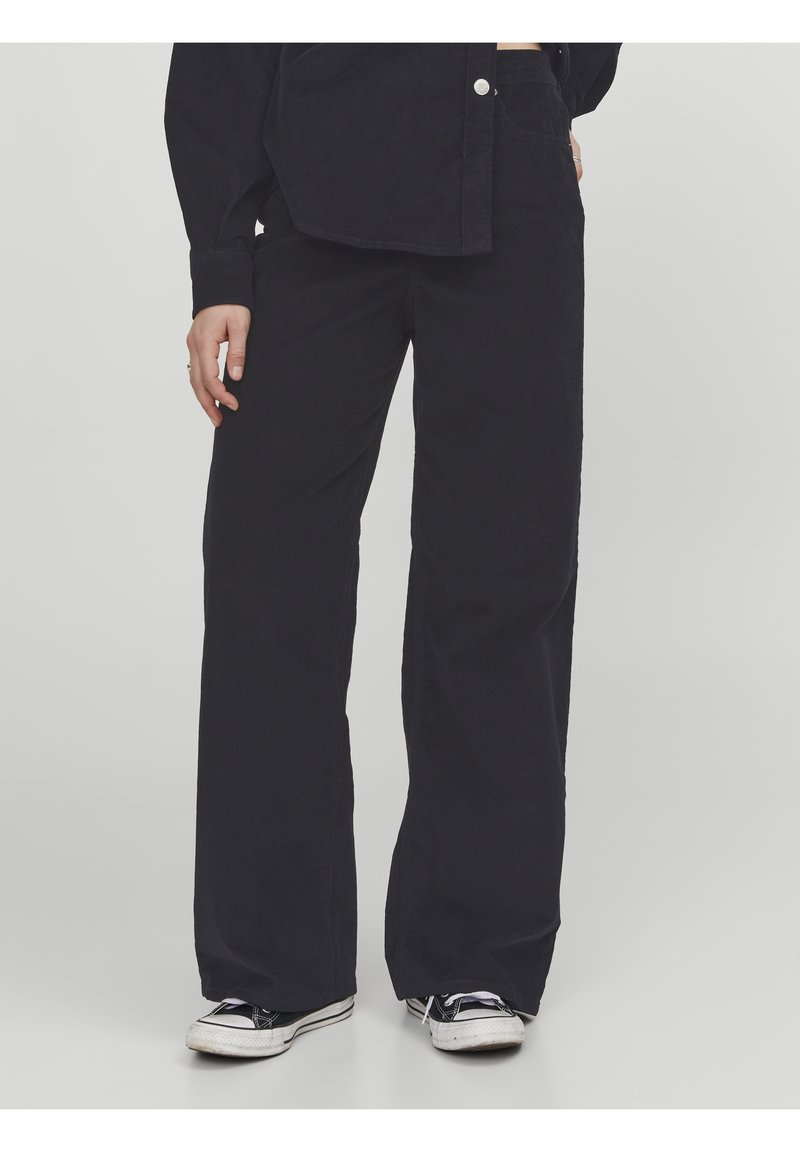 Personne portant un pantalon noir ample, une chemise noire à boutons et des baskets noir et blanc, debout sur un fond gris uni.