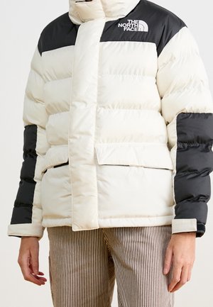 Cream and black The North Face puffer jacket with front pockets worn over brown striped pants on a standing person.
