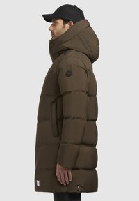 Braune Pufferjacke mit hohem Kragen und Kapuze, ausgestattet mit einer gesteppten Textur, Seitentaschen und einem schwarzen Logo-Patch am Ärmel.