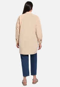 Camicia beige a maniche lunghe con orlo dritto e polsini abbottonati; abbinata a jeans in denim blu e sandali chiari. Tessuto dalla superficie liscia.