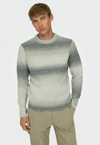 Maglione a gradiente grigio con collo rotondo e polsini a coste. Il tessuto appare morbido con una trama a maglia media, abbinato a pantaloni di velluto a coste beige.