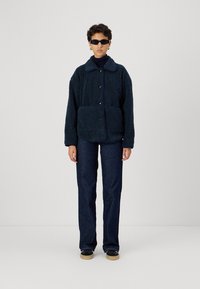 Giacca in pile blu navy con tessuto texturizzato, ampio colletto, bottoni frontali e tasche laterali, abbinata a jeans scuri e scarpe nere.