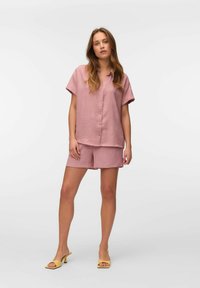 Camisa de botón de manga corta rosa y pantalones cortos a juego, hechos de un tejido texturizado. Combinados con sandalias de tacón abiertas amarillas.