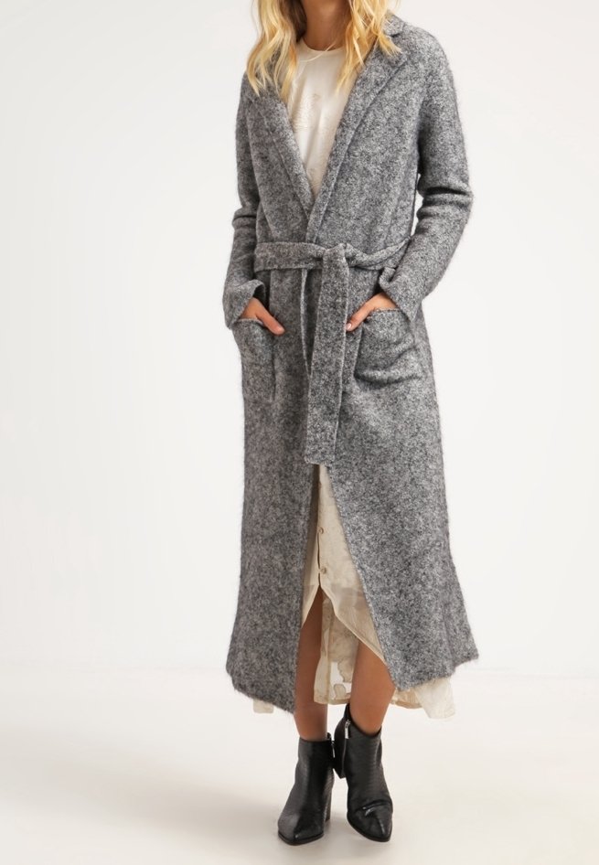 Femme portant un long cardigan en tricot gris ceinturé sur une robe claire et des bottines noires, debout les mains dans les poches.