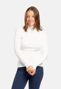 Top bianco a maniche lunghe con colletto alto realizzato in tessuto morbido, design aderente con forellini per il pollice, abbinato a jeans blu scuro.