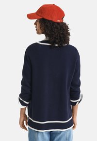 Navy sweater with white trim, featuring long sleeves and a relaxed fit. Paired with a bright red corduroy cap. Casual style.
