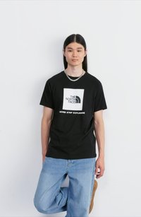 Camiseta de algodón negra con un logotipo blanco cuadrado que incluye el texto "THE NORTH FACE" y "NEVER STOP EXPLORING". Lleva puesta unos jeans de color azul claro.