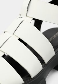 Sandal blanc avec plusieurs larges brides, fabriqué en matériau lisse, avec une semelle intérieure noire moulée et une semelle texturée pour une meilleure adhérence.