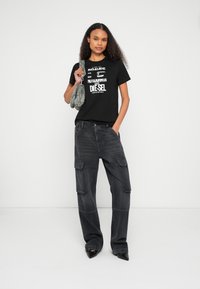 Čierne bavlnené tričko s bielym grafickým textom, kombinované s voľnými tmavosivými cargo nohavicami s vreckami a čiernymi špicatými podpätkami. Drží vzorovanú tašku.